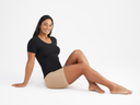 A woamn is wearing the Holly Short Sleeve Top in black with Shapewear Shorts in almond while sitting on the floor with arms stretched behind her. Yummie