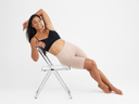 A woman is wearing the Rosie in Almond with a black bra while reclining on a chair. Yummie
