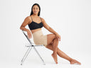 A woman is wearing the Audrey Bra in Black and the Brigitta Mid Waist Shapewear Slip Short - Yummie SoftServe™ in ALmond while sitting in a chair with one leg extended and the other bent. Yummie