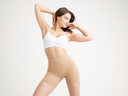 A woman is wearing the Ultralight Shapewear Short in Almond with a white bra standing with her arms at side and hands in hair. Yummie