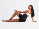 A woman is wearing the Rosie Shapewear Slip Short - 360Sculpt™ Cotton in Black with a white bra while reclining on the ground. Yummie