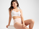 A woman is wearing the Convertible Scoop Bralette in White with the Non-Shaping Retro Brief in Rose Smoke while sitting. Yummie