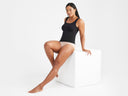 A woman is wearing the Anna Scoop Neck Shaping Tank Top - 360Sculpt™ Cotton in black with briefs in Almond while sitting on a white block. Yummie