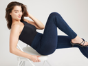 A woman is wearing the Denim Shaping Legging in True Indigo with a one shoulder bodysuit in black while leaning back in a folding chair. Yummie