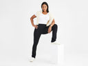 A woman is wearing the Jodi Slim Bootcut Shaping Legging - 360Sculpt™ Cotton in black with a white top and cream sweater over shoulders while standing with one leg on a column. Yummie