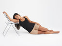 A woman is wearing the Fay Convertible Zoned Shapewear Camisole in black with a shapewear short in almond while reclining on the ground. Yummie