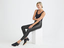 A woman is wearing the Faux Leather Legging w/ Zip in black with a black tank Top while sitting on a column. Yummie
